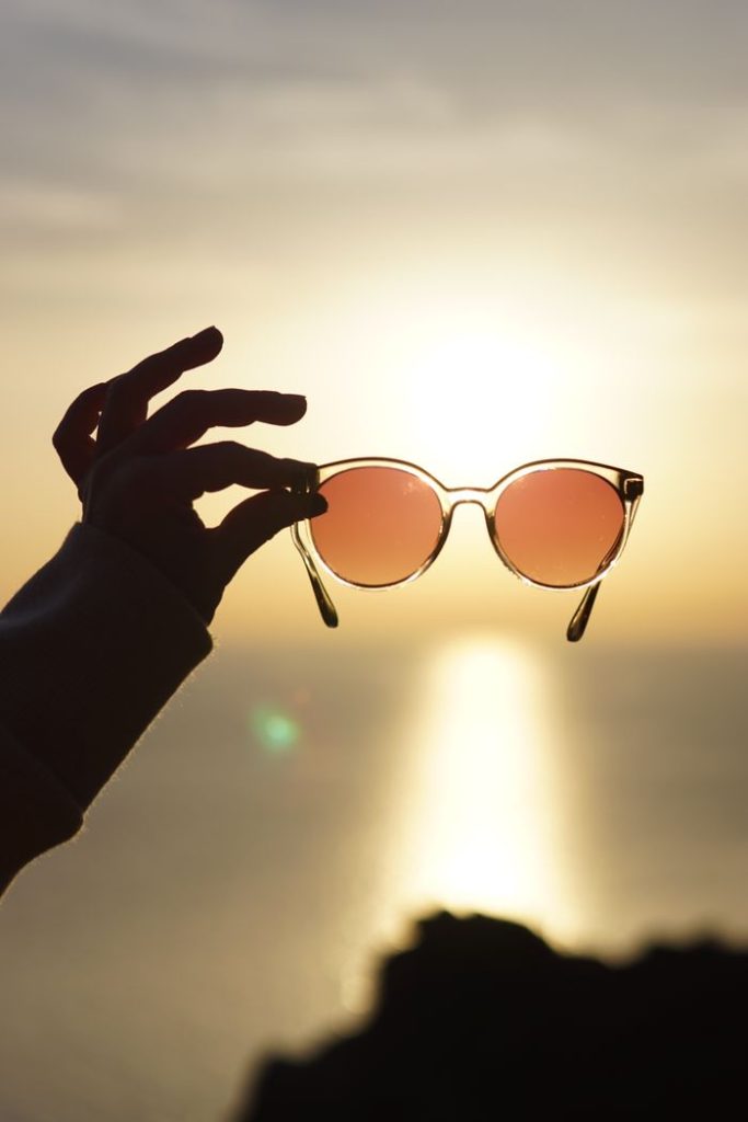 A hand holding sunglasses with a sunset view over the water, creating a serene golden hour silhouette.