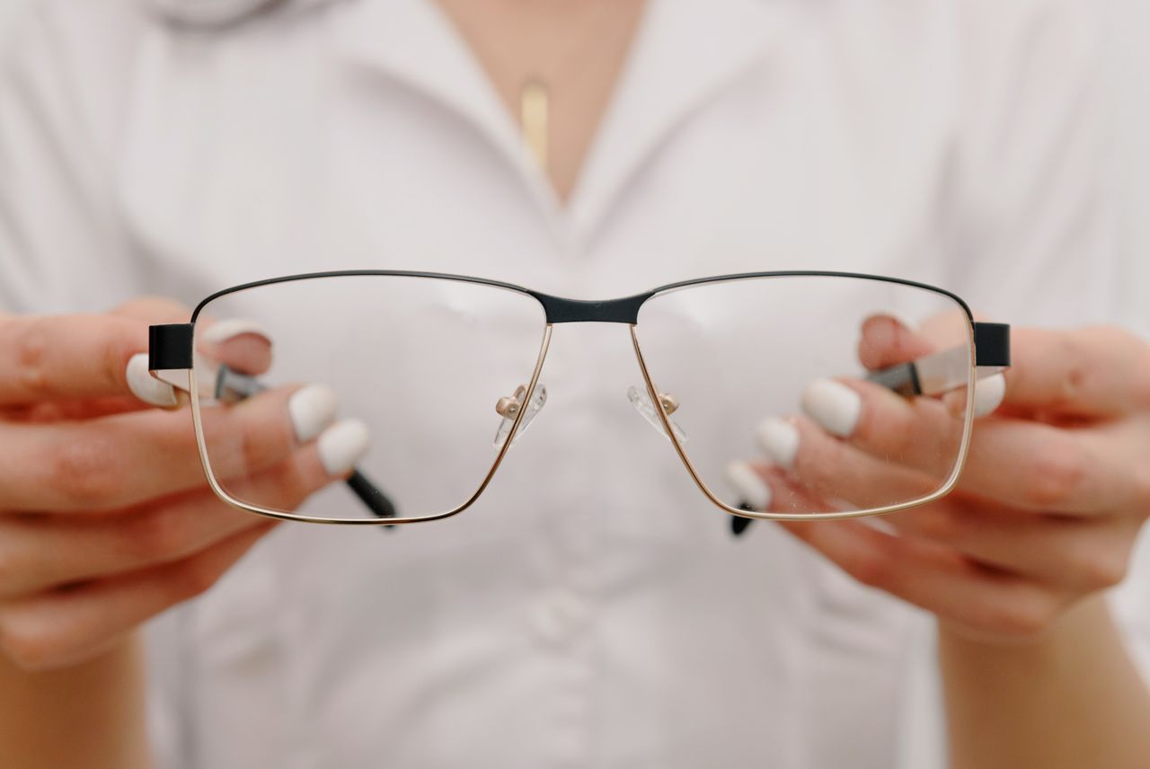 services-03 Crop anonymous female with manicured hands wearing white robe and showing classic eyeglasses in light modern optometrist clinic